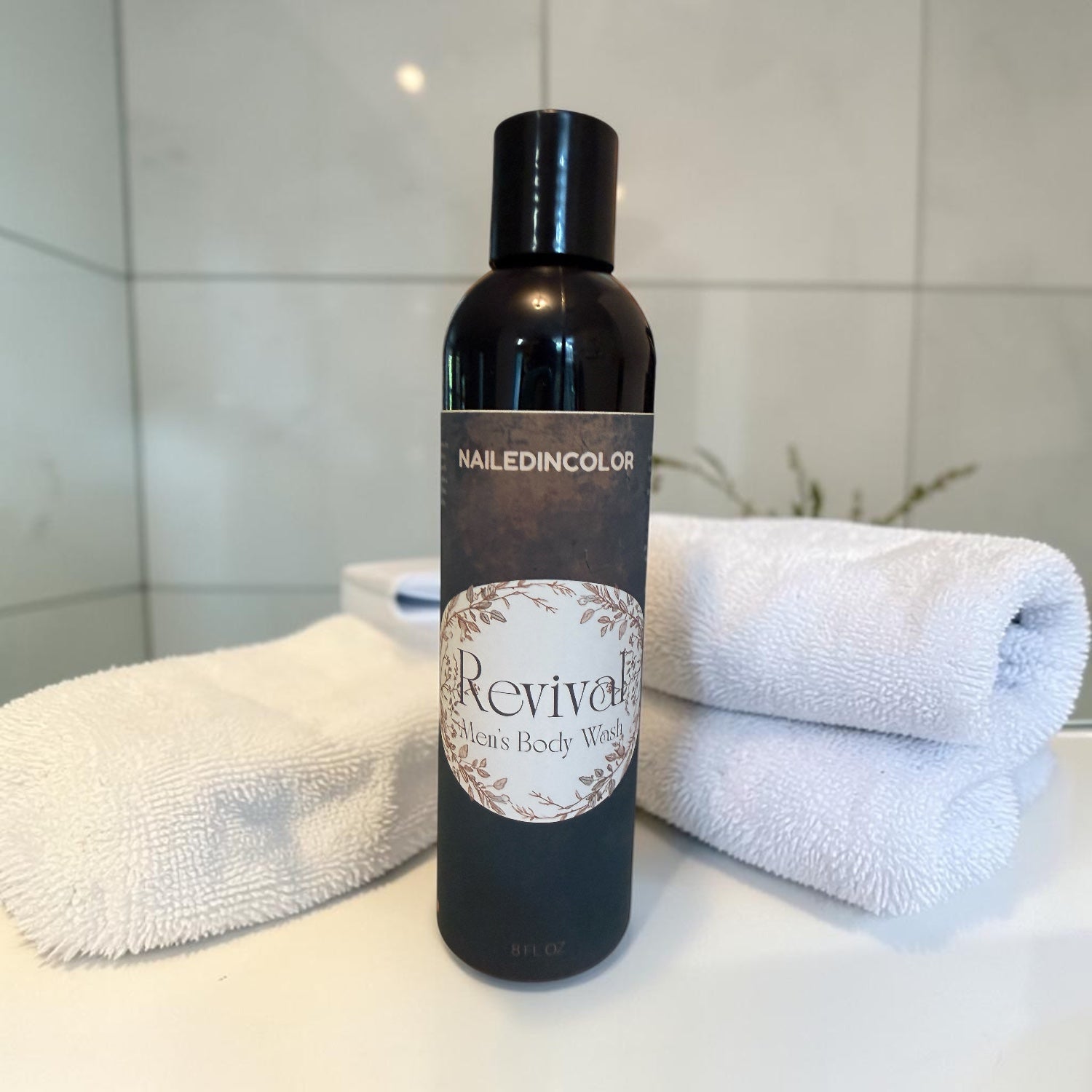 A bottle of Revival Cleansing Body Wash by Nailed In Color stands on a white surface with folded white towels behind it against a light tiled wall.
