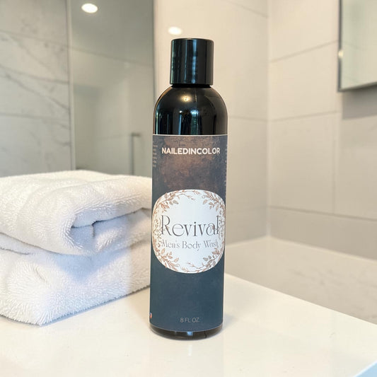 A bottle of NailedinColor Revival Body Wash stands on a white bathroom counter next to folded white towels, with a softly blurred tiled bathroom background.