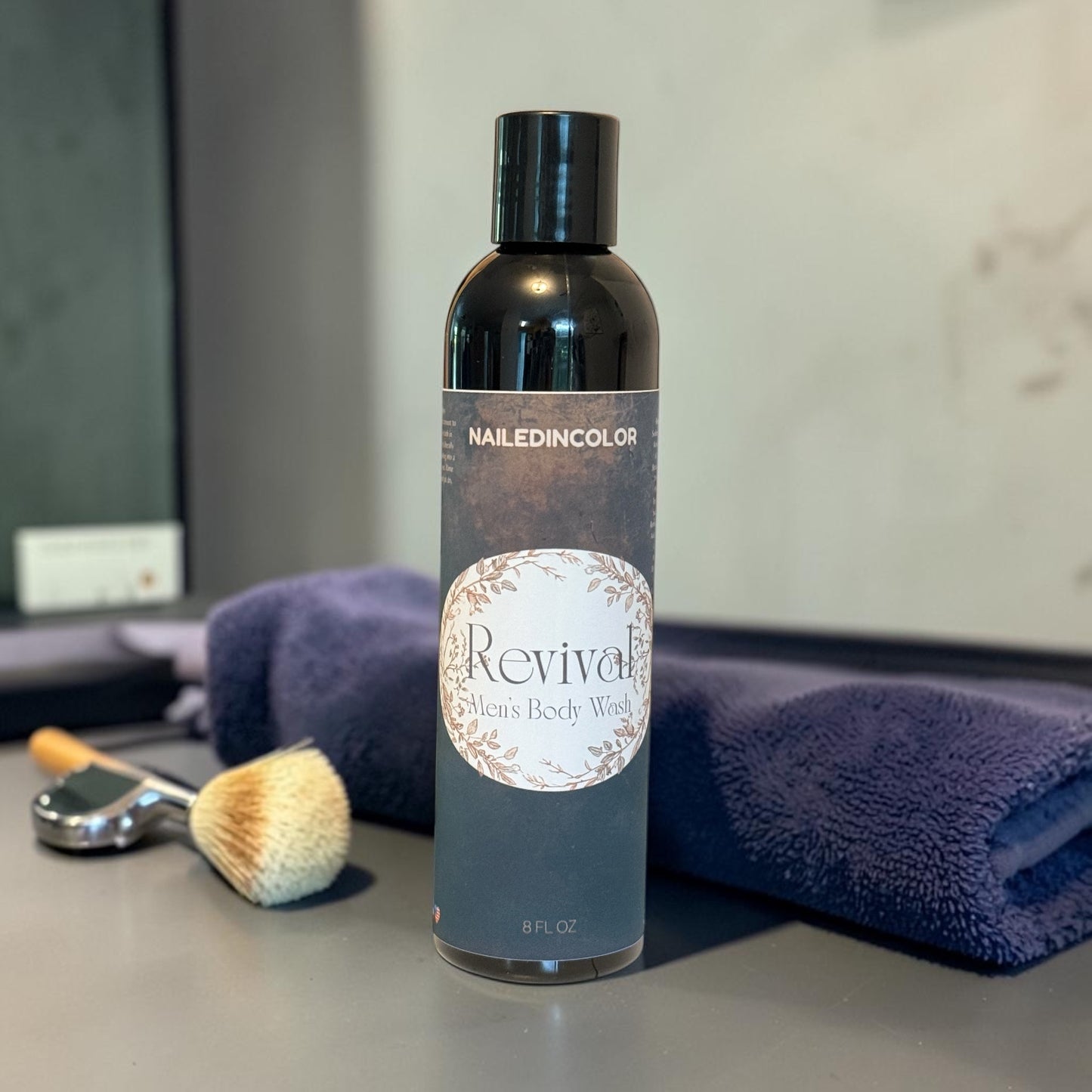A bottle of NailedinColor Revival Body Wash stands on a countertop with a folded blue towel and a small shave brush behind it. The bottle is dark with a black cap and a gray label featuring a white oval label