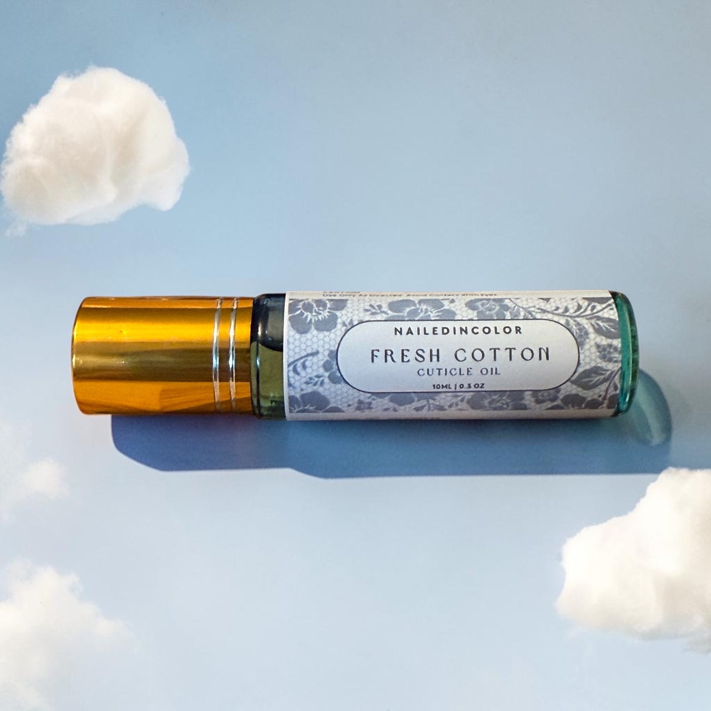 A 10ml roller bottle of Nailedincolor Fresh Cotton Cuticle Oil, featuring a metallic gold cap and a white patterned label, set against a light blue background with stylized white clouds.