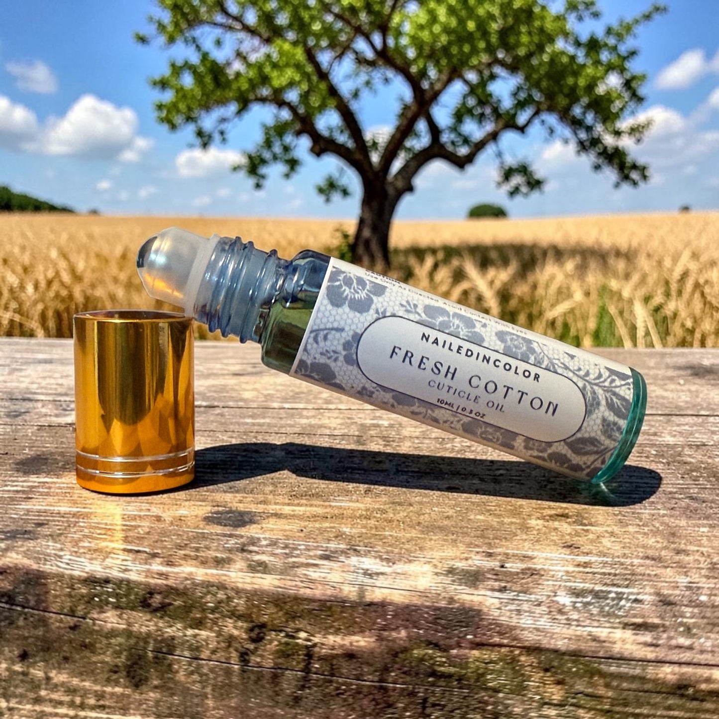 A bottle of NailedinColor Fresh Cotton Cuticle Oil lies on its side on a weathered wooden surface outdoors, with its gold cap placed beside it. A blurred tree and golden field are visible in the background under a blue sky.