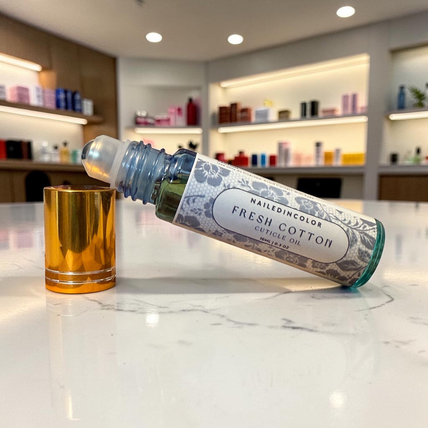 A bottle of NailedinColor Fresh Cotton Cuticle Oil lies on its side on a white marble countertop, with its gold cap placed beside it. The background shows a softly blurred beauty salon or retail interior with shelves and lighting.