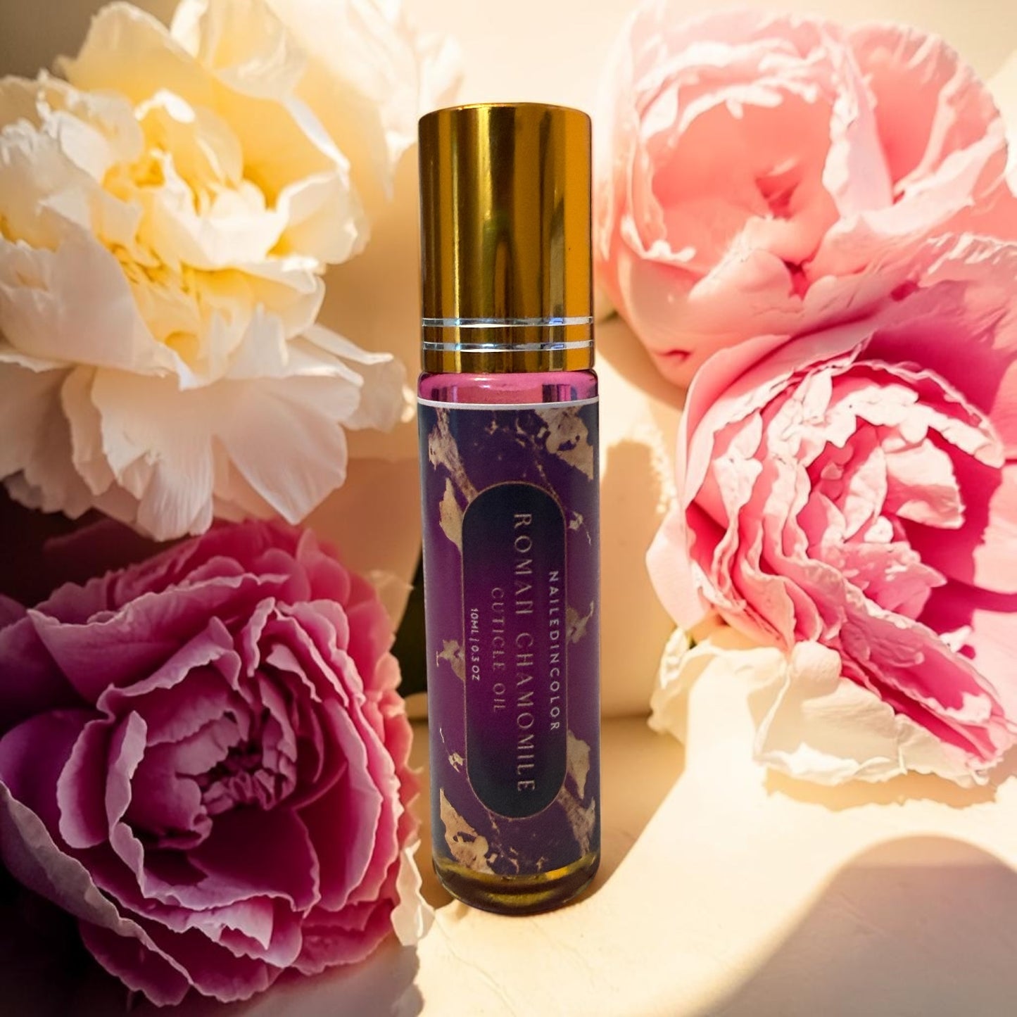 A bottle of Roman Chamomile cuticle oil stands upright in the center, surrounded by large pink, magenta, and cream peony flowers on a warm beige surface