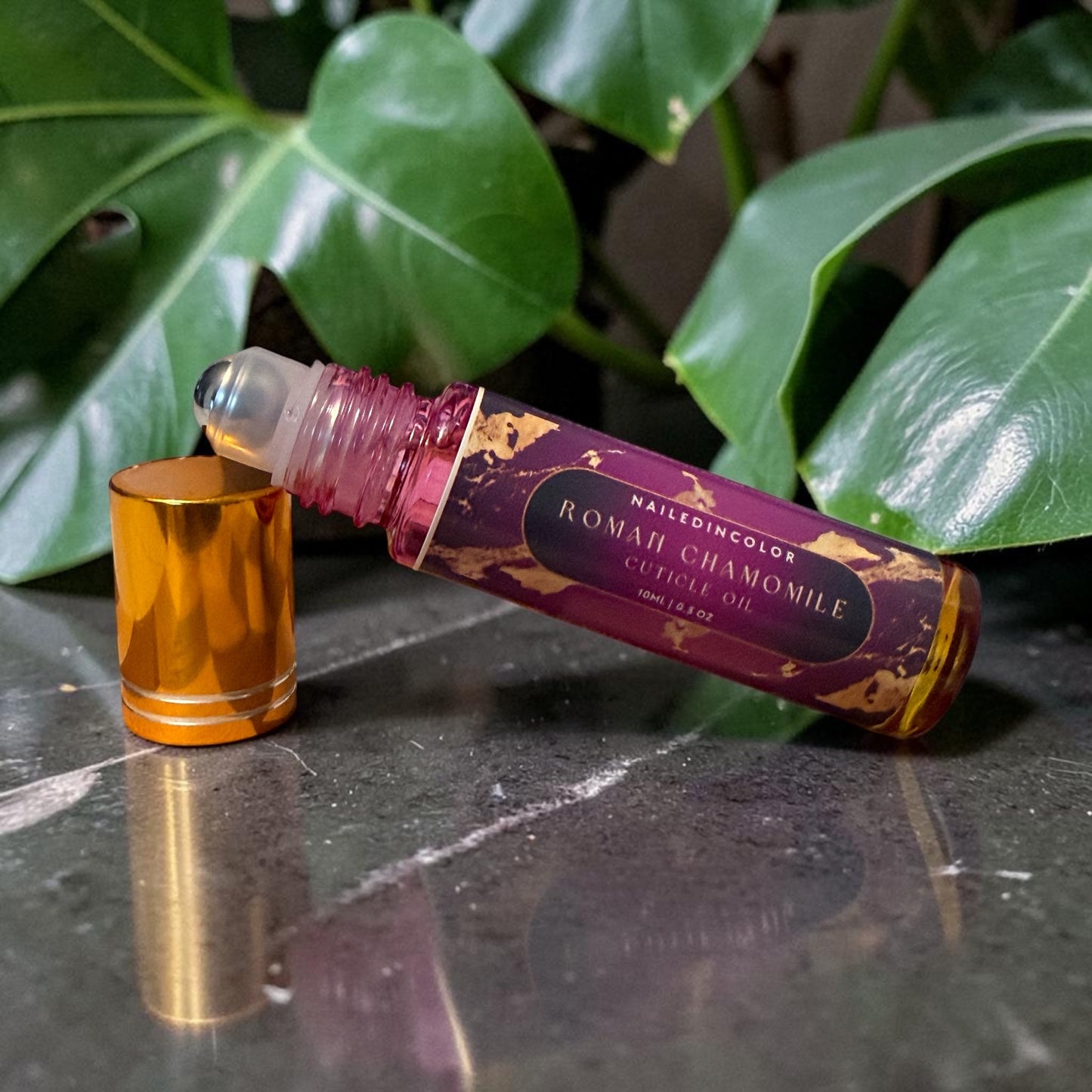 A roll-on bottle of Roman Chamomile  Oil with its cap removed, resting on a dark marble surface with green leaves in the background.