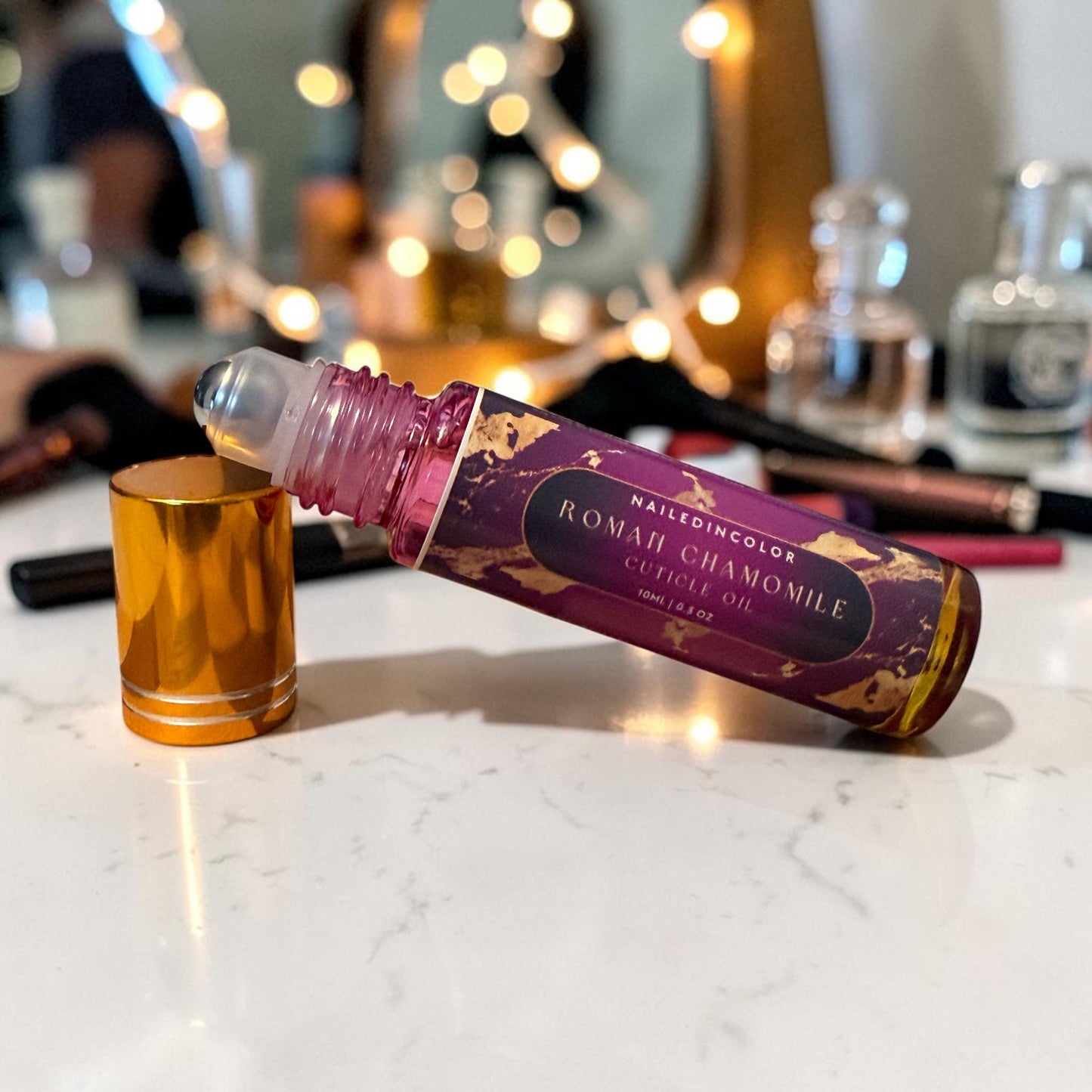 A bottle of  Roman Chamomile  Oil roll-on, with its metallic gold cap removed and placed beside it on a white marble surface. The background is a softly blurred vanity setting with warm bokeh lights and cosmetic items.