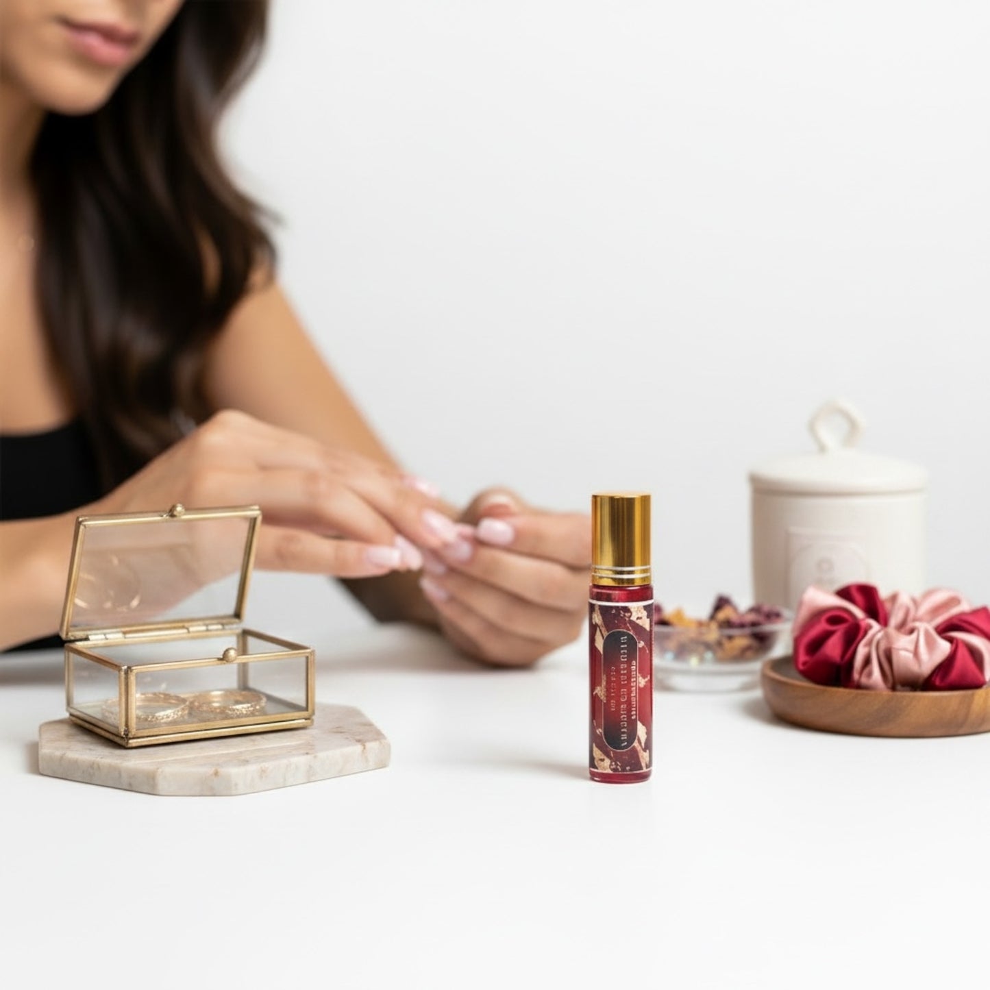 A woman sits at a white table with her hands visible, next to a small gold-capped cuticle bottle with a red label. On the table are also a clear glass jewelry box with gold trim, a white ceramic jar, and two pink velvet scrunchies.