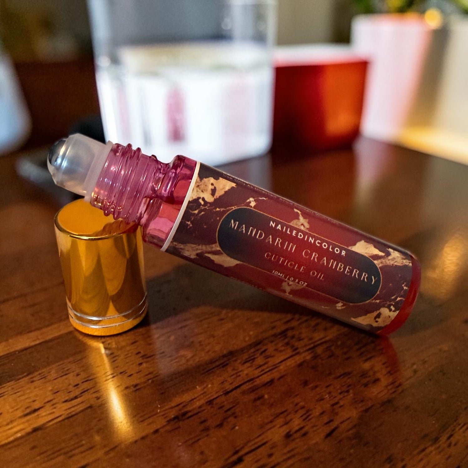 A cuticle oil rollerball bottle labeled Mandarin Cranberry Cuticle Oil' resting on a wooden table next to its gold cap.