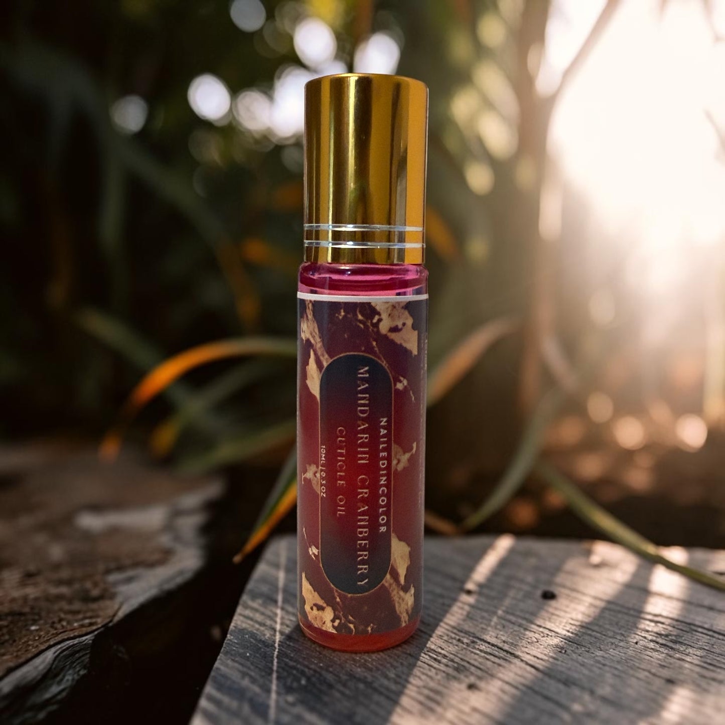A small cylindrical bottle of Mandarin Cranberry cuticle oil with a shiny gold cap, standing on a weathered wooden surface with a blurred outdoor garden background and bright sunlight.