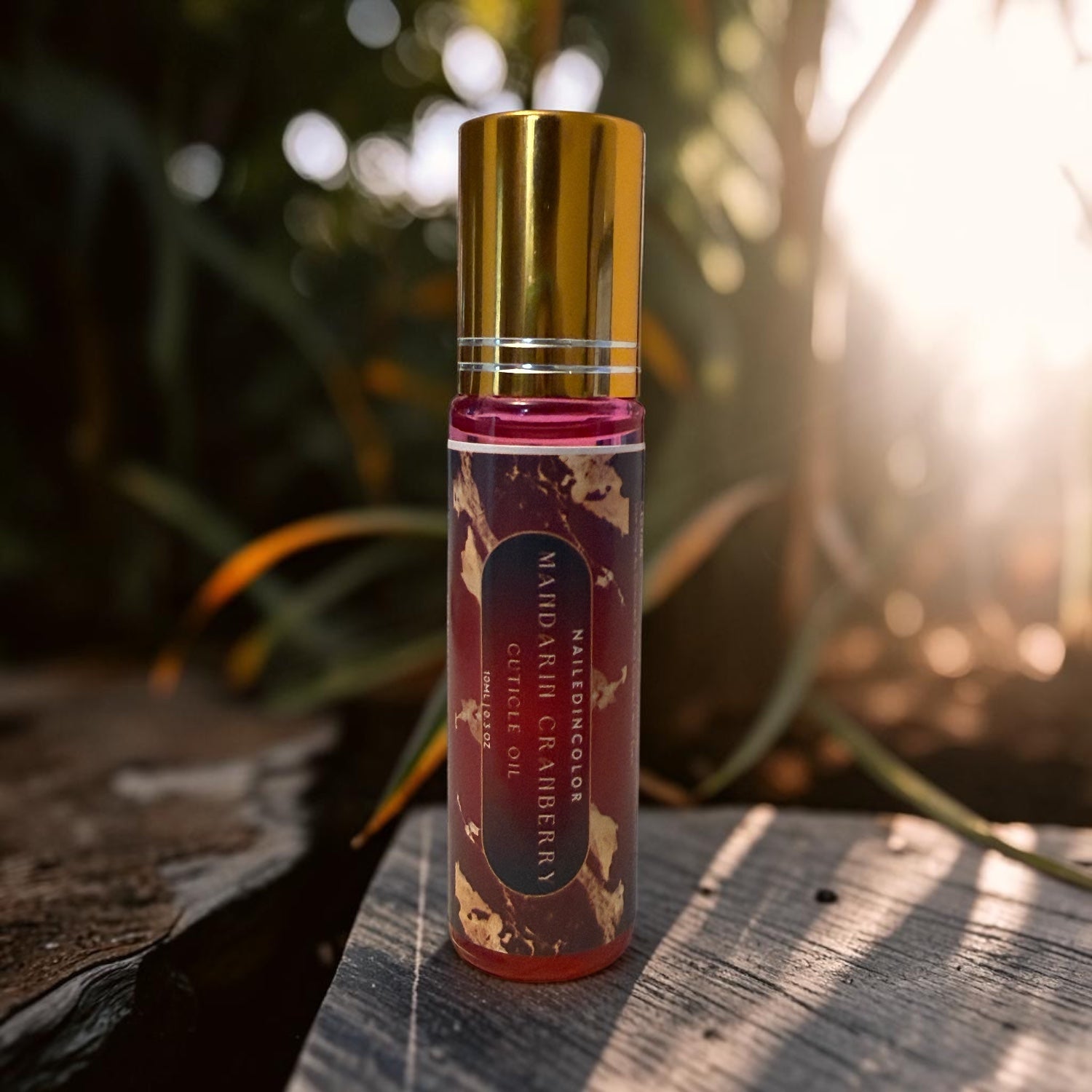 A small cylindrical bottle of Mandarin Cranberry cuticle oil with a shiny gold cap, standing on a weathered wooden surface with a blurred outdoor garden background and bright sunlight.
