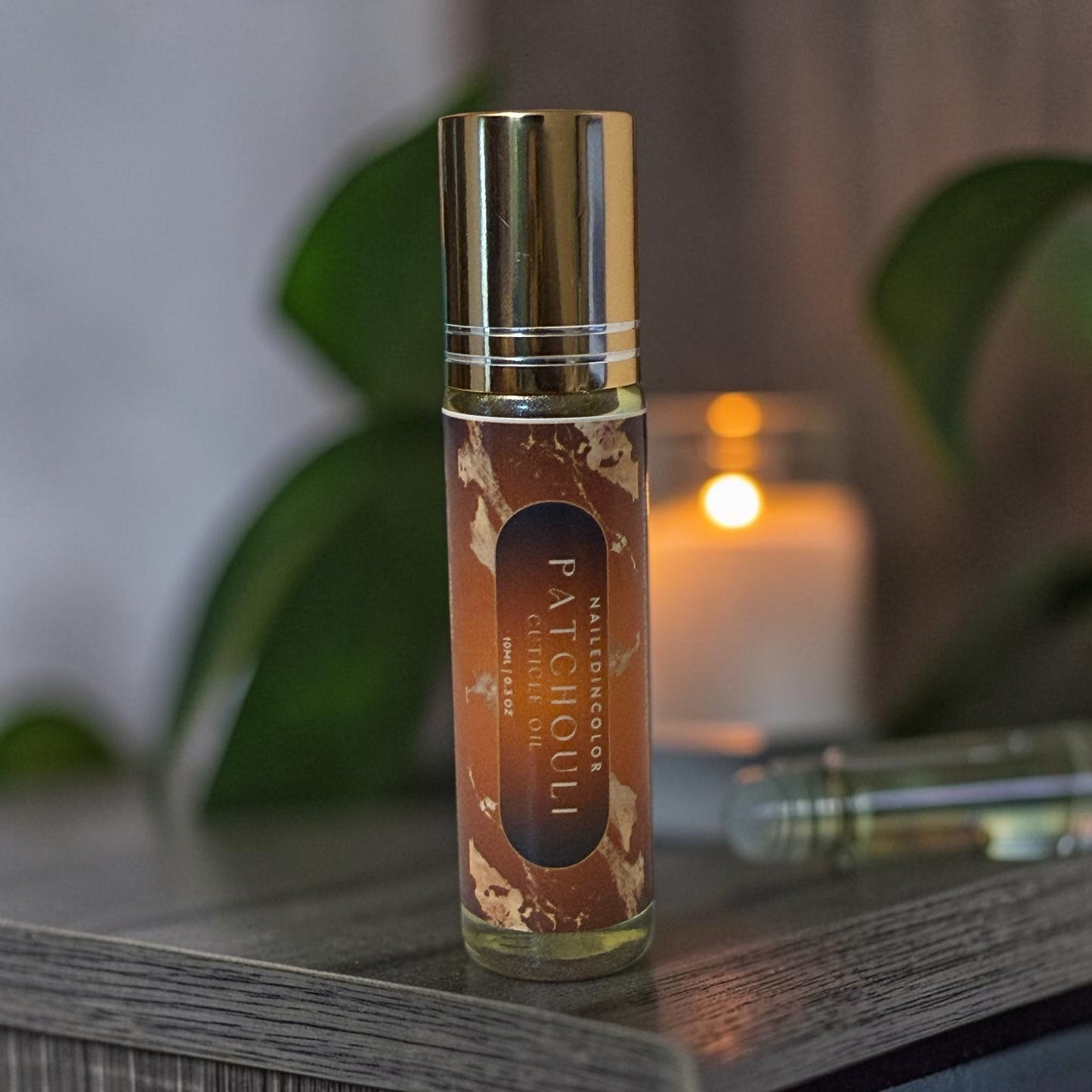 A roll-on bottle of Patchouli  cuticle oil with a gold cap and brown label, standing on a dark wooden surface with a blurred candle and green leaves in the background.