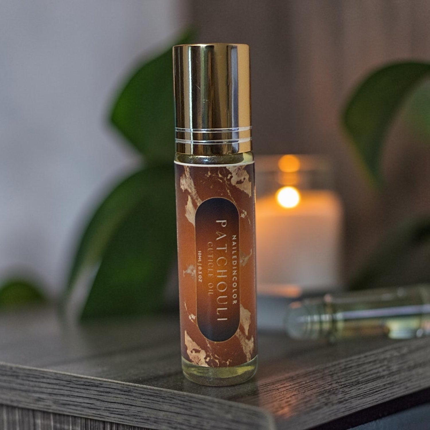 A roll-on bottle of Patchouli  cuticle oil with a gold cap and brown label, standing on a dark wooden surface with a blurred candle and green leaves in the background.