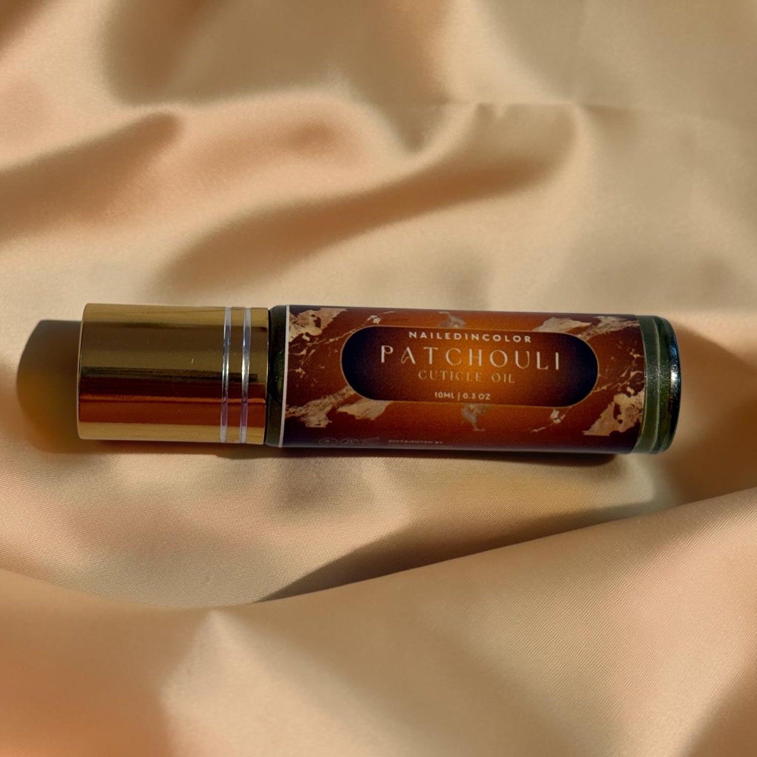 A roll-on bottle of Nailedinicolor Patchouli Cuticle Oil with a metallic gold cap and a brown patterned label, resting on draped beige satin fabric.