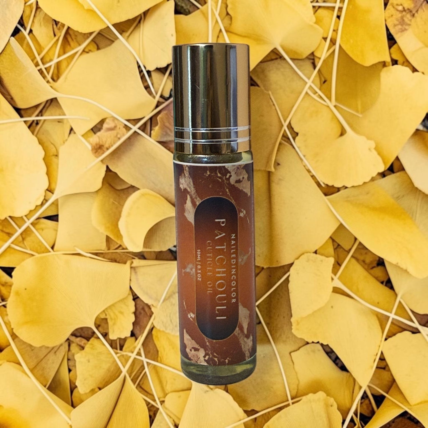 A cylindrical glass roller bottle of cuticle oil, featuring a reflective silver cap and a label reading 'Patchouli'  placed on a bed of yellow ginkgo leaves.