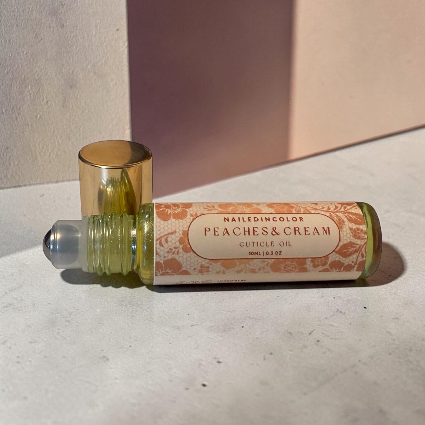 A roll-on bottle of cuticle oil labeled 'NAILEDINCOLOR PEACHES & CREAM CUTICLE OIL' with a gold cap, resting on a textured light gray surface with a pink background panel.