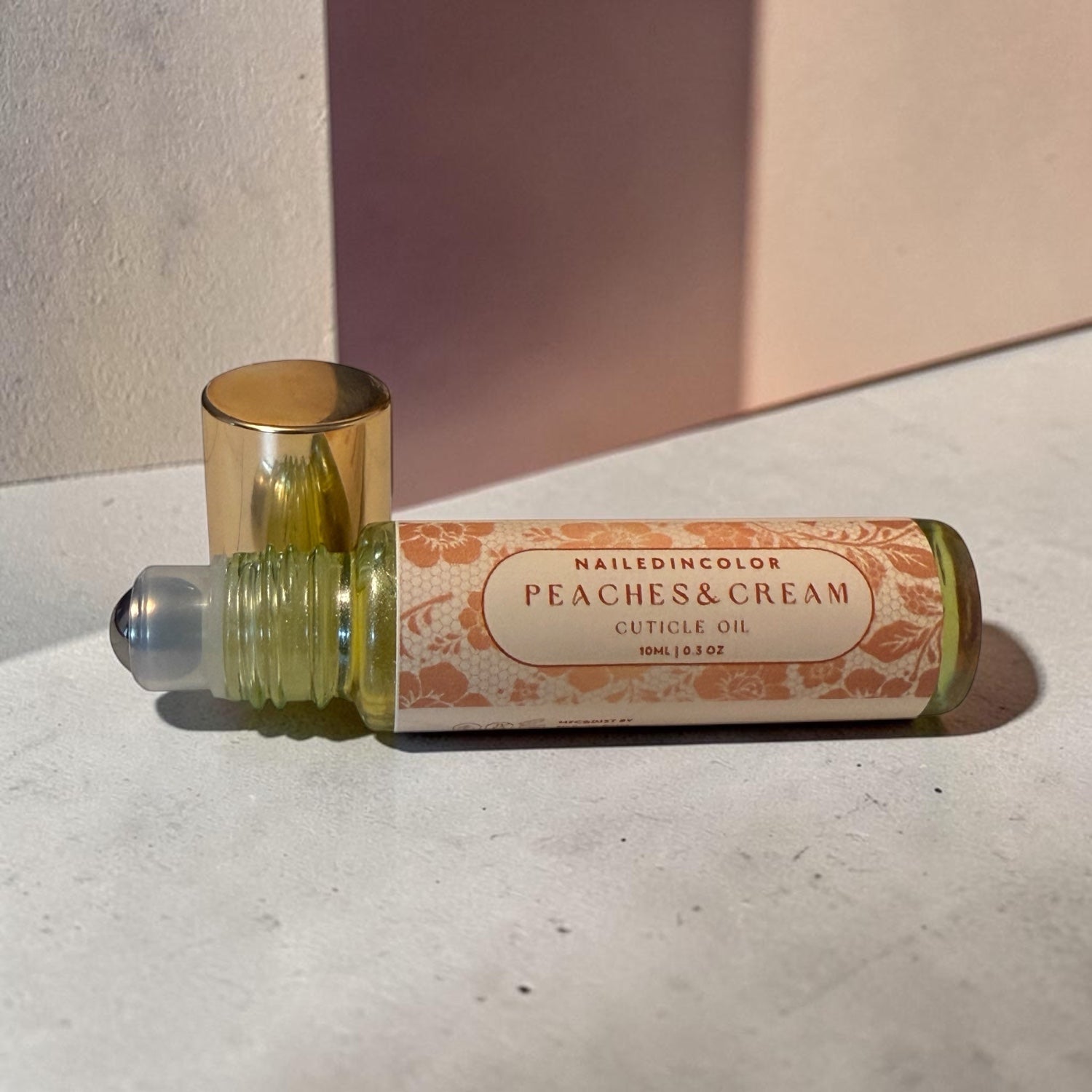 A roll-on bottle of cuticle oil labeled 'NAILEDINCOLOR PEACHES & CREAM CUTICLE OIL' with a gold cap, resting on a textured light gray surface with a pink background panel.