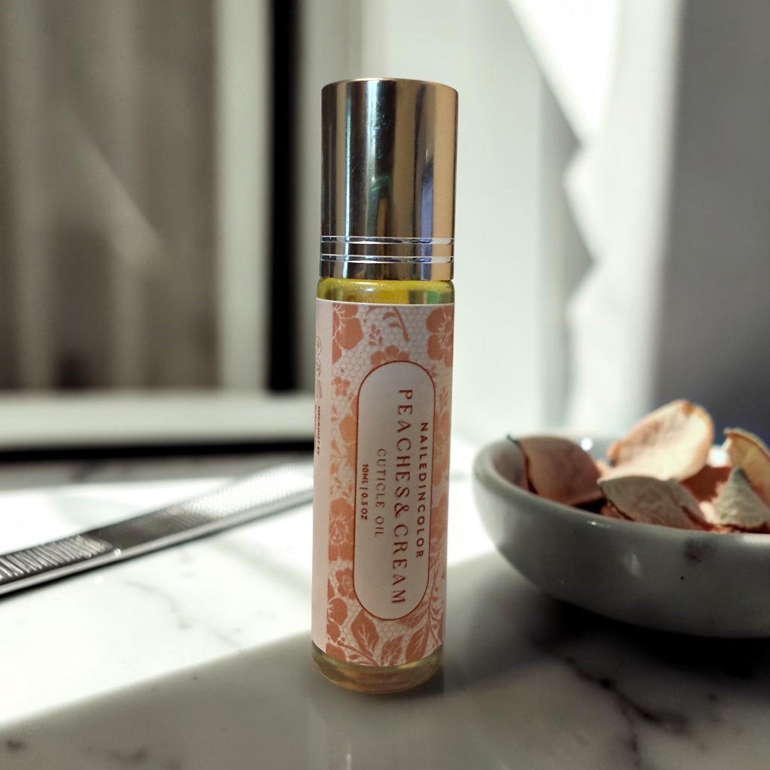 A small roller bottle labeled 'NailedInColor PEACHES & CREAM' cuticle oil stands on a marble surface. The bottle has a shiny silver cap and a peach-toned label with floral detailing.
