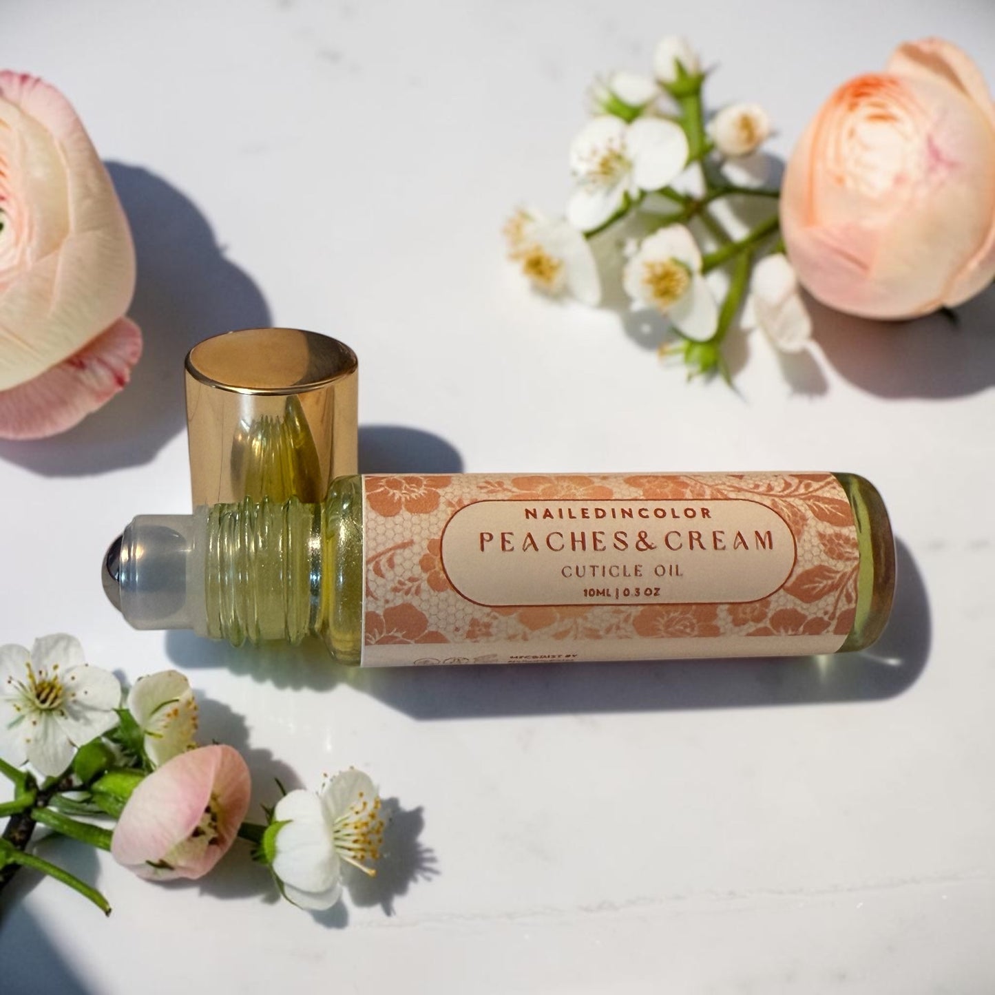 A cuticle oil roll-on bottle with a gold cap and a peach-colored label reading 'NAILEDINCOLOR PEACHES&CREAM CUTICLE OIL 10ML (0.3 OZ)', styled on a white surface with pink roses and white blossoms.