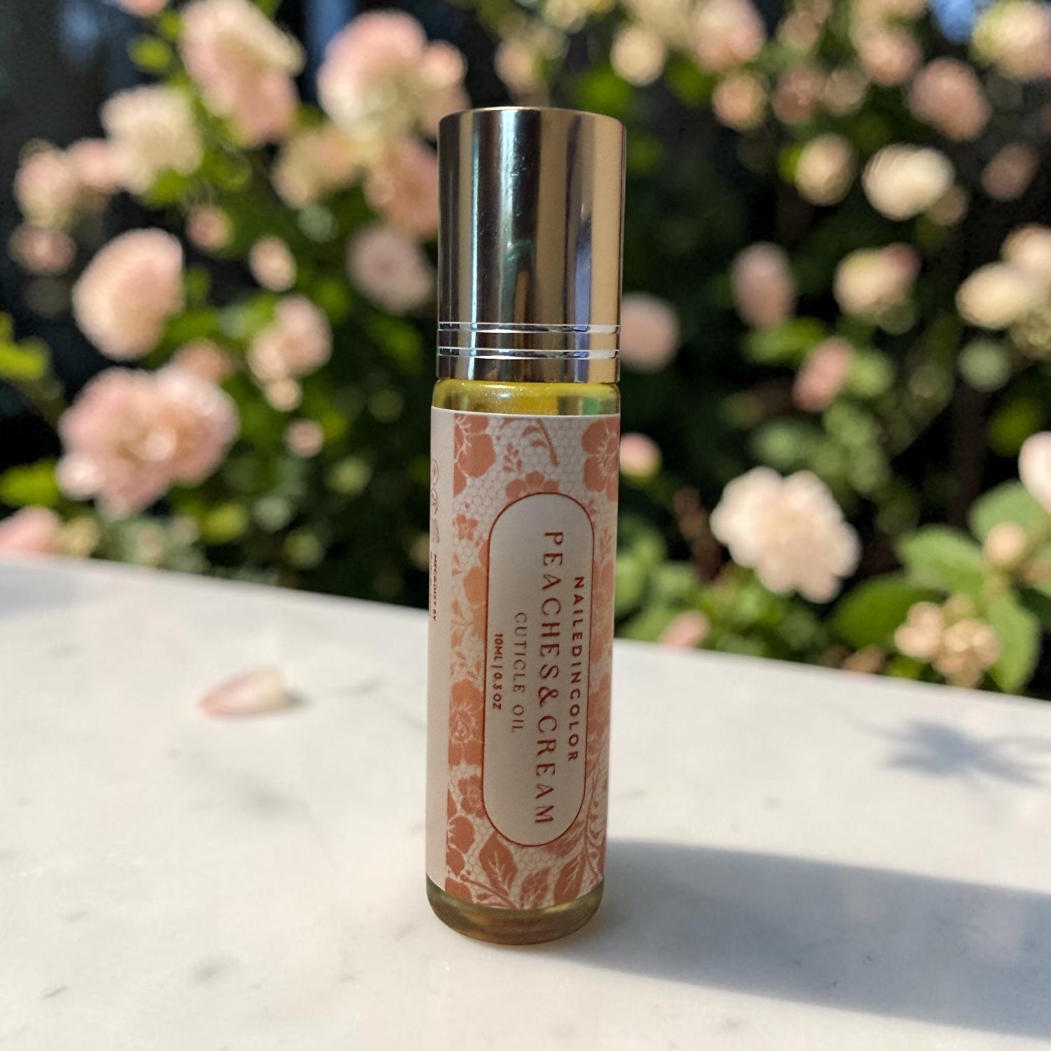 A small cylindrical cosmetic bottle labeled 'NailedInColor Peaches & CREAM' with a shiny silver cap, standing on a light stone surface with a blurred background of pink flowers and green foliage.