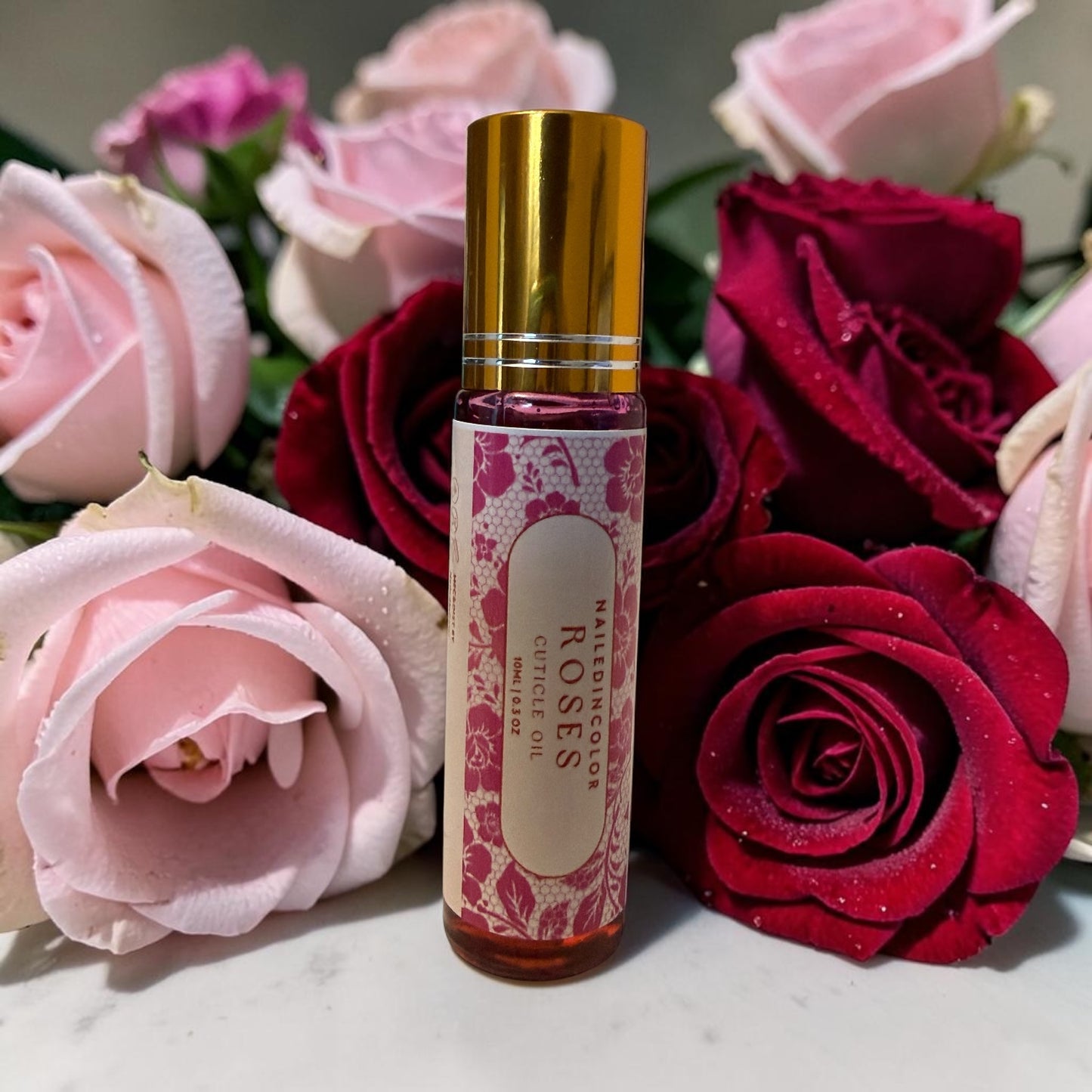 A small cylindrical roller bottle labeled 'ROSES' with a gold cap, standing upright among pink and deep red roses on a light marble-like surface.