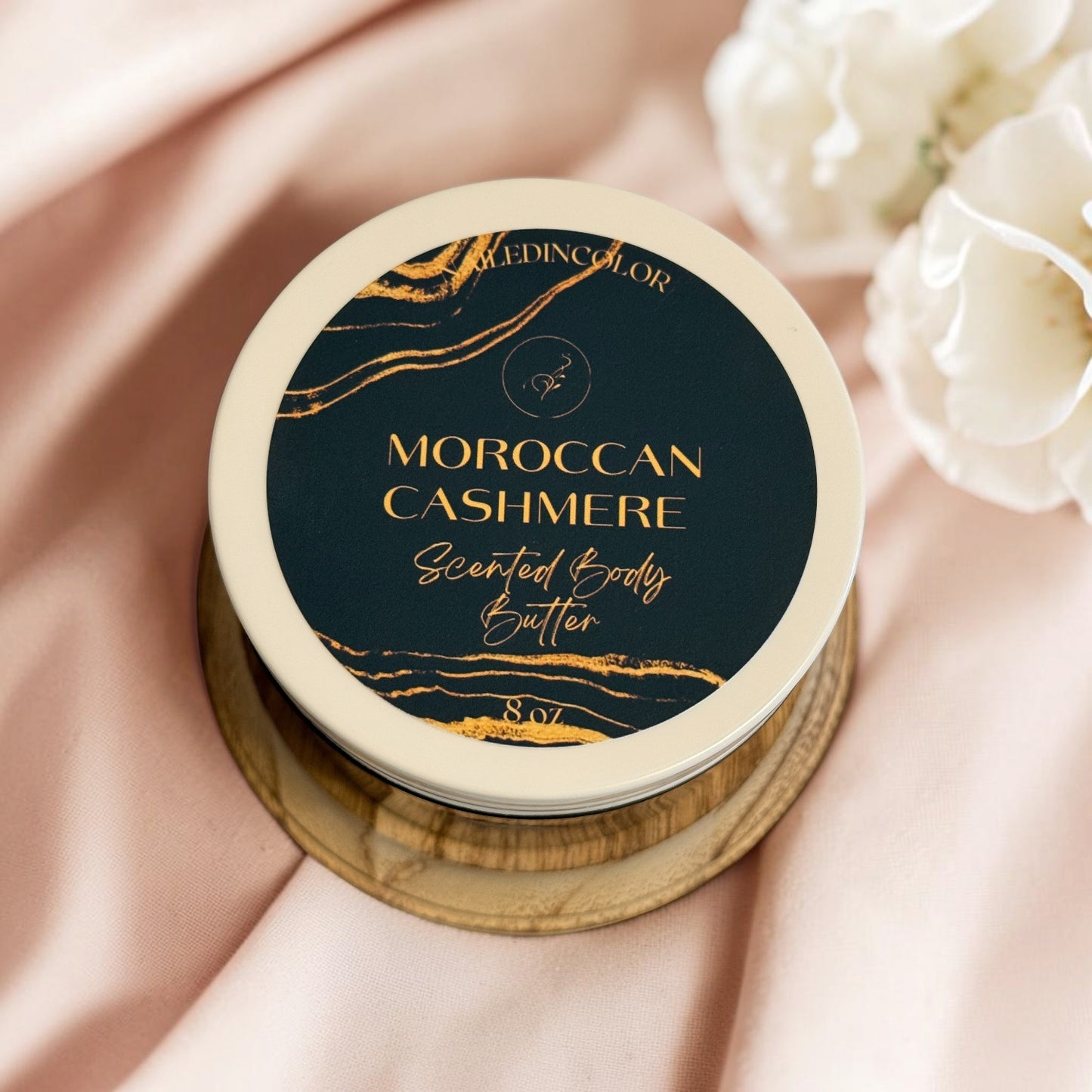 A jar of Moroccan Cashmere Scented Body Butter on a wooden stand resting on soft pink fabric, blur of white lace in background.