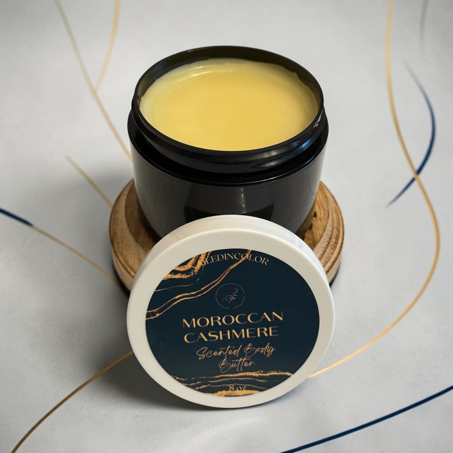 A jar of Moroccan Cashmere Scented Body Butter with its lid placed in front, set on a small wooden coaster against a light gray background with thin gold and blue decorative lines.