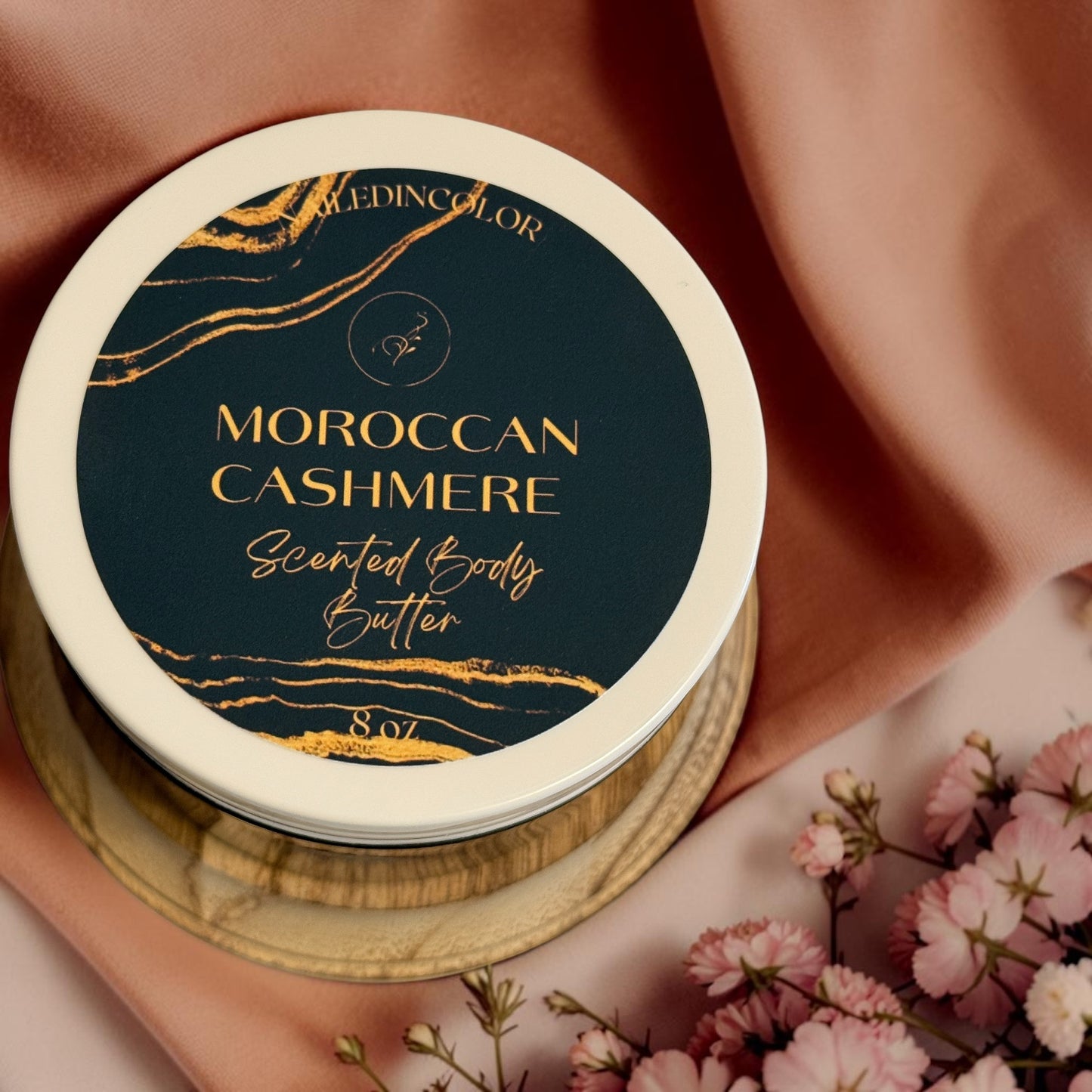 A round jar of NailedInColor Moroccan Cashmere Scented Body Butter, featuring a dark label with gold lettering and gold marbled accents. The jar is stacked on a gold-colored wooden coaster, set against a draped dusty pink fabric backdrop with pink flowers in the foreground.
