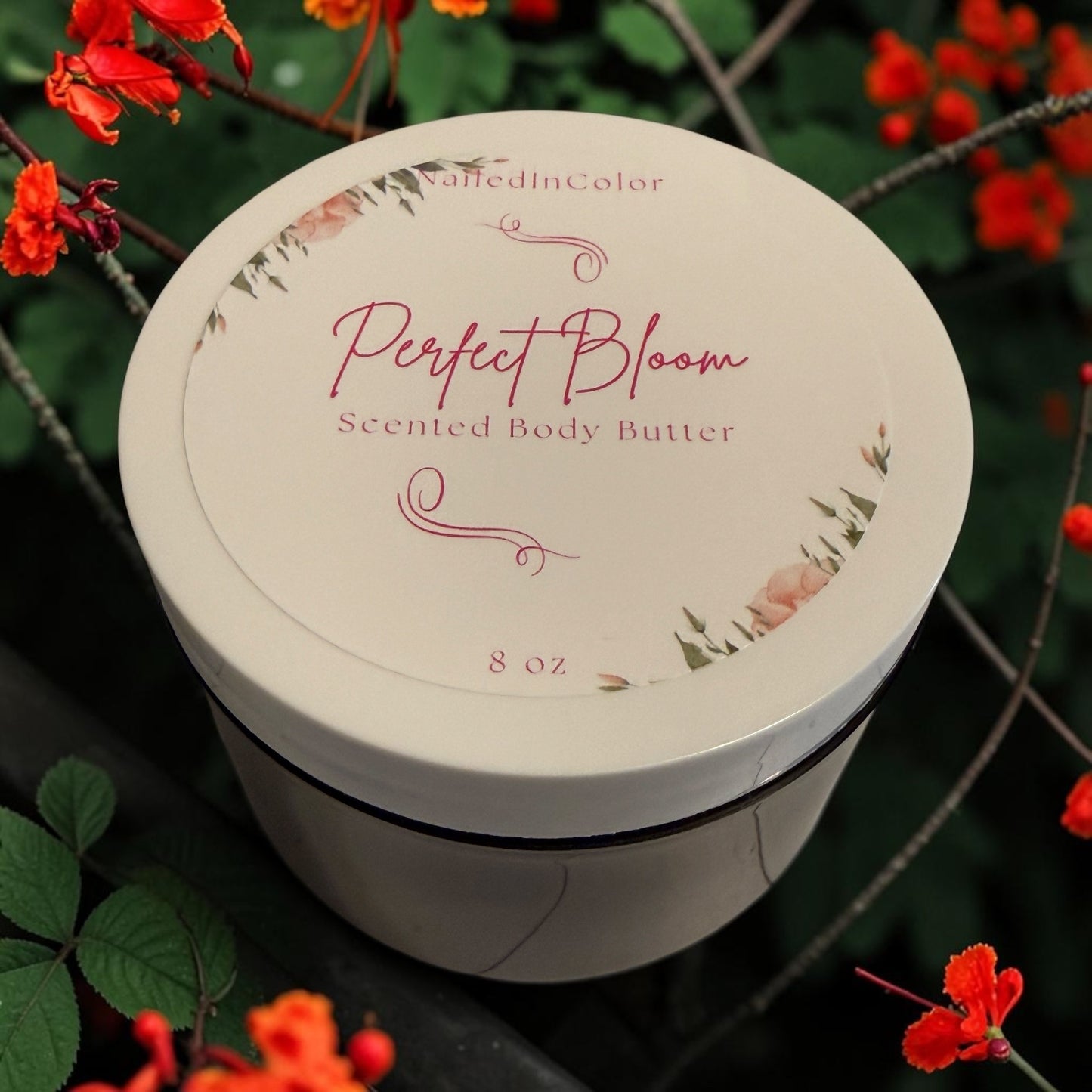 A round white jar of 'Perfect Bloom' Scented Body Butter by NailedInColor is displayed outdoors among green leaves and bright red flowers. The lid features elegant pink script and floral illustrations.