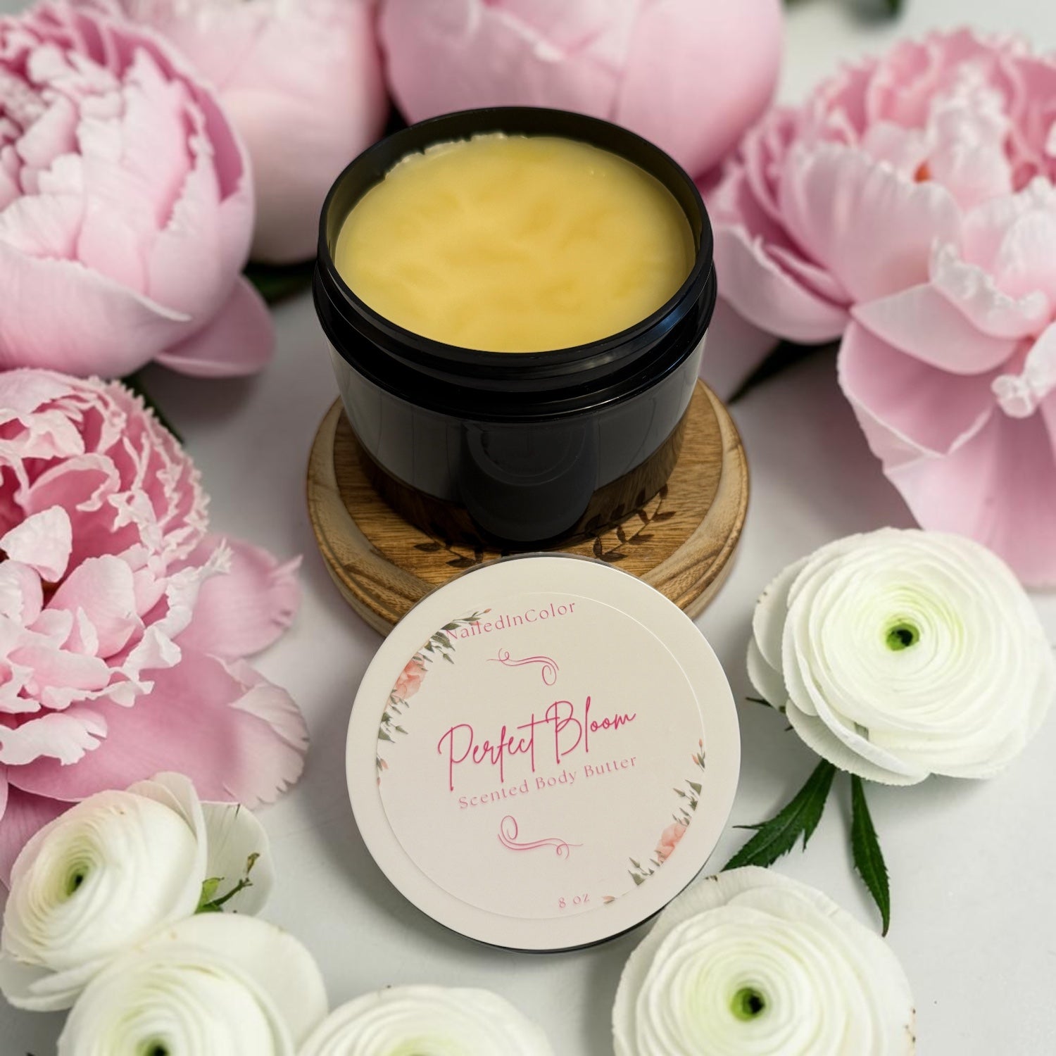 A black jar of NailedInColor Perfect Bloom Scented Body Butter filled with pale yellow balm, displayed on a wooden coaster with its floral-patterned lid in front. The jar is surrounded by pink peonies and white ranunculus flowers on a white surface.