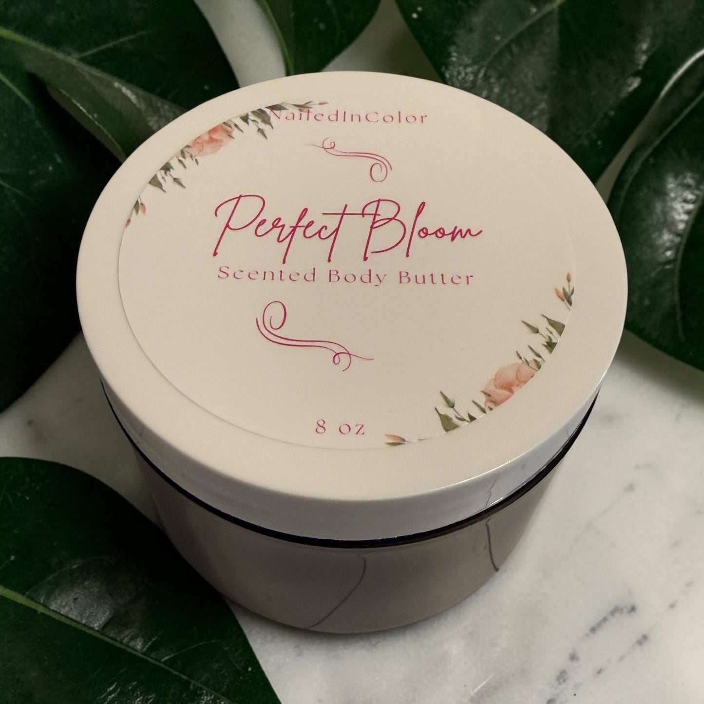 A round jar of NailedInColor Perfect Bloom Scented Body Butter, featuring a cream-colored lid with pink floral illustrations and pink script lettering, set on a white marble surface surrounded by green leaves.