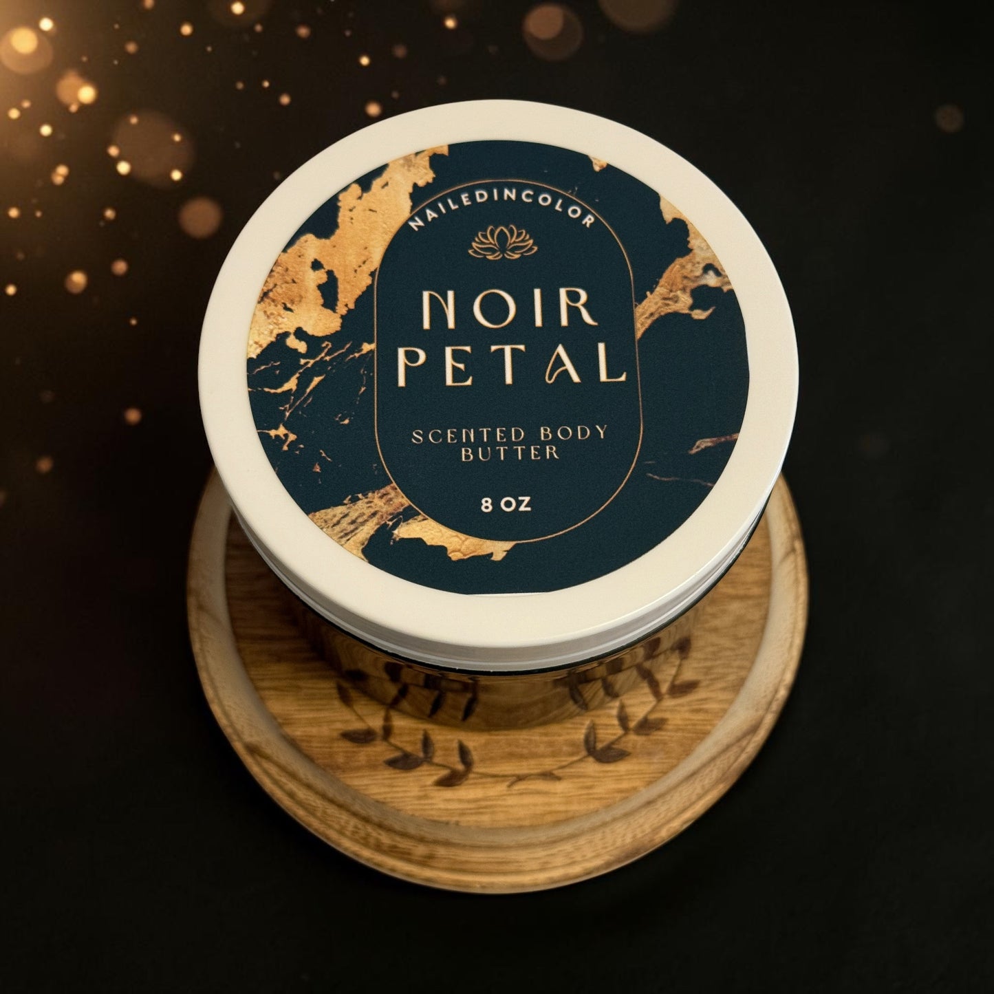 A round jar of Nailed In Color Noir Petal Scented Body Butter sits on a carved wooden stand against a dark background with warm golden bokeh lights. The lid features a navy blue label with gold marbled accents and gold lettering.