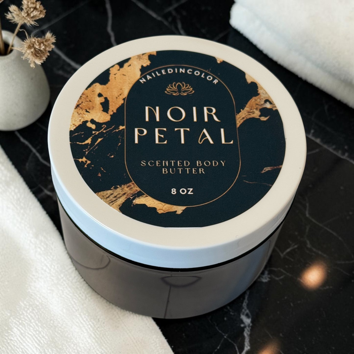 An 8 oz jar of NailedinColor Noir Petal Scented Body Butter, featuring a dark navy and gold marbled lid, set on a black marble surface with white towels and small white vase with field flowers