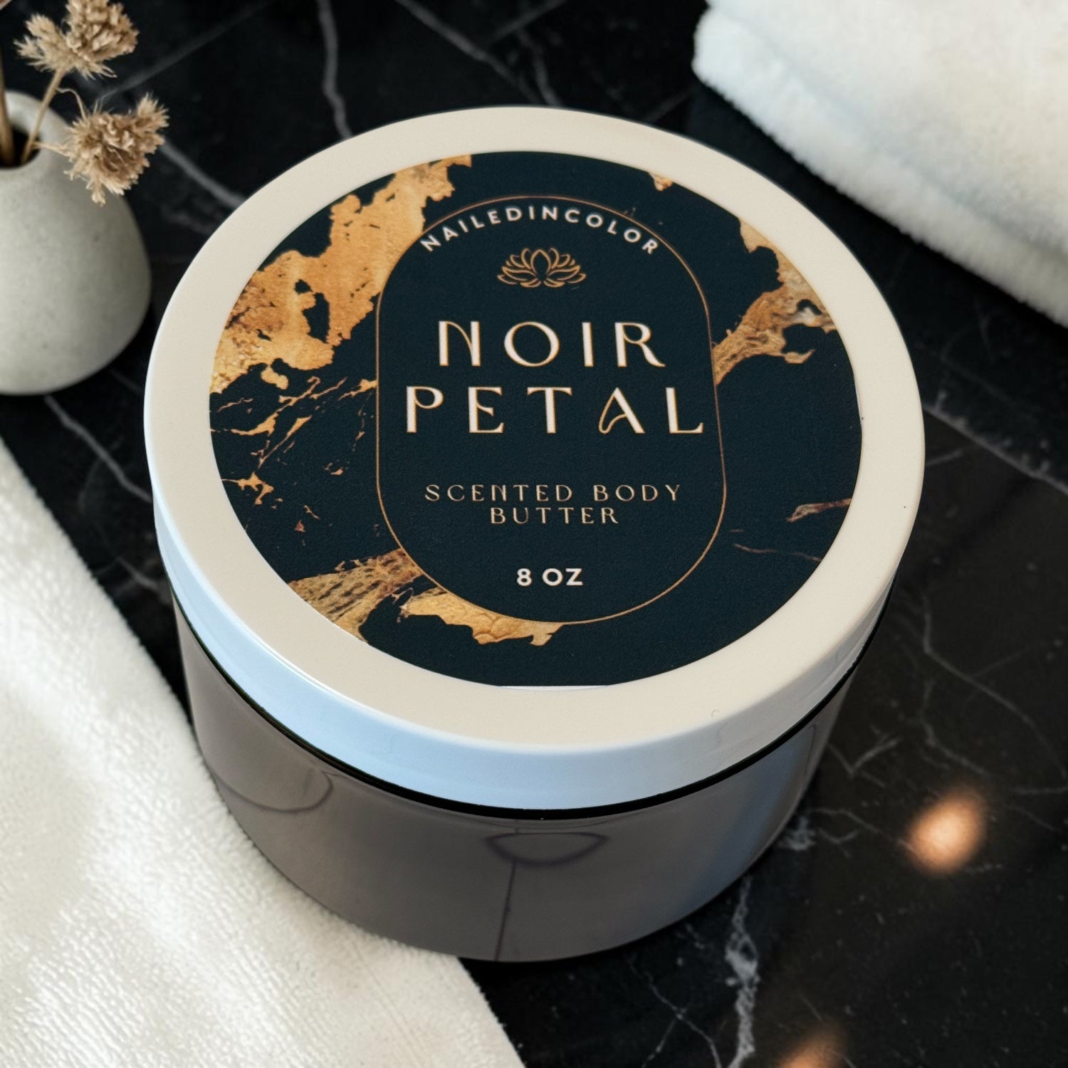 An 8 oz jar of NailedinColor Noir Petal Scented Body Butter, featuring a dark navy and gold marbled lid, set on a black marble surface with white towels and small white vase with field flowers
