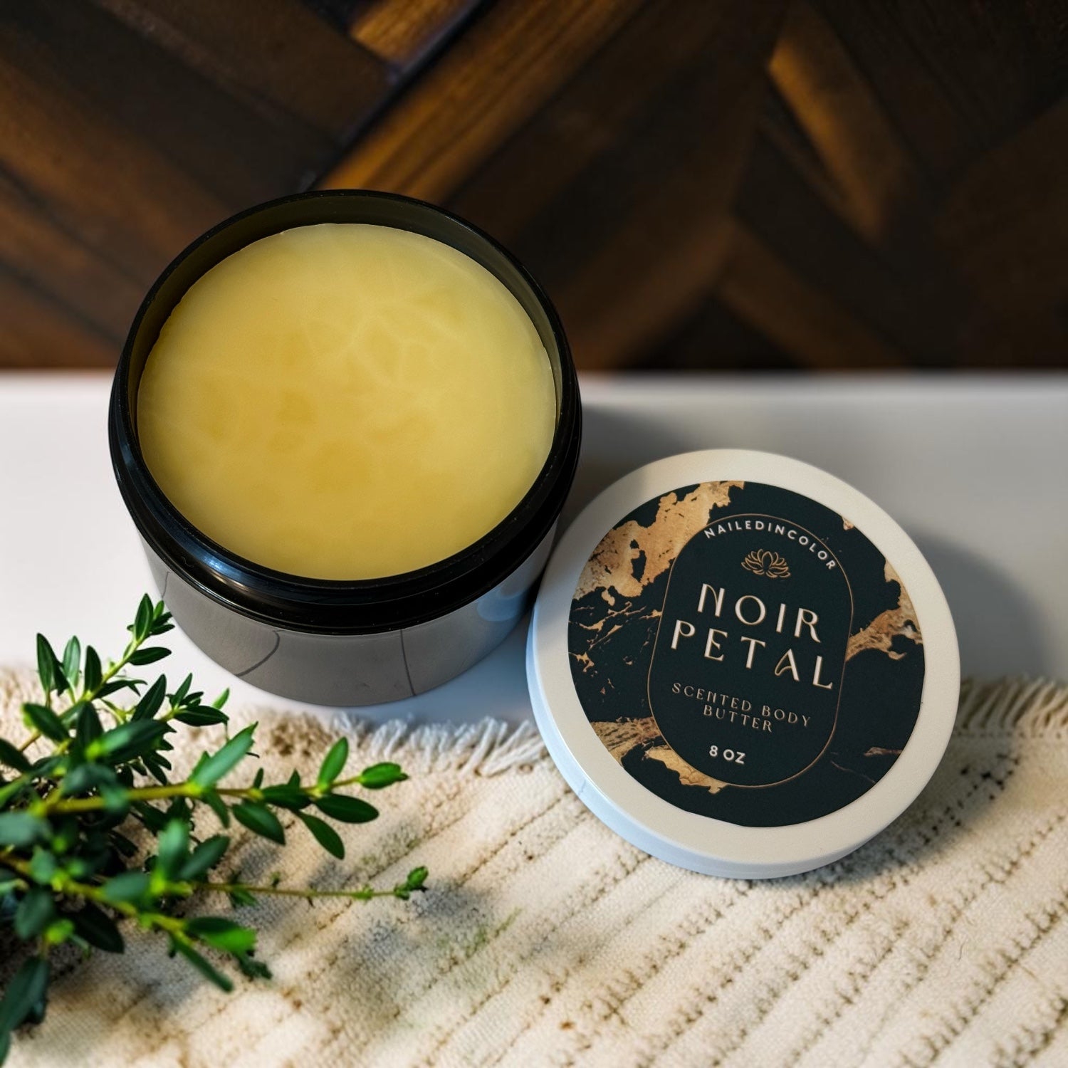 An open jar of Noir Petal, cap sits next to jar on a white placemat with greenery and in background is wood parquet