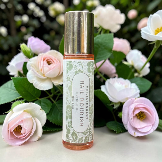 A bottle of Nail Nourish cuticle oil by NailedInColor stands centered on a light surface, surrounded by pink and white roses with green leaves in an outdoor garden setting.