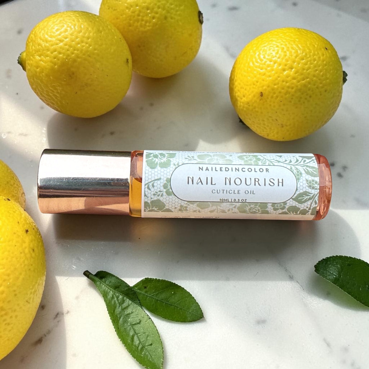 A roll-on bottle of NailedinColor Nail Nourish Cuticle Oil lies on a white marble surface, surrounded by whole lemons and green leaves. The bottle has a white label with green floral patterns and a metallic rose gold cap.