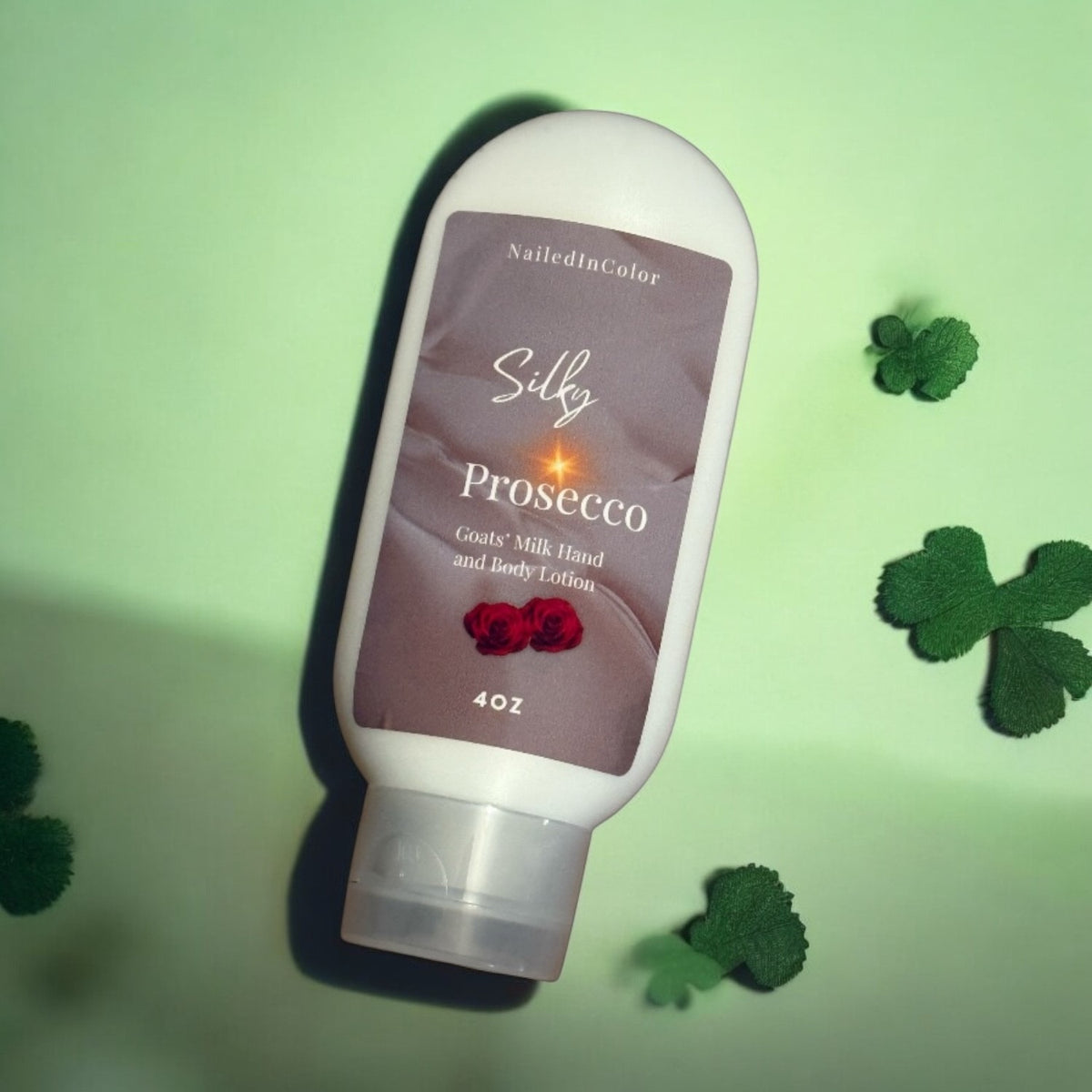 A 4 oz bottle of NailedInColor Silky Prosecco Goats Milk Hand and Body Lotion, displayed diagonally on a light green background with decorative clover leaves.