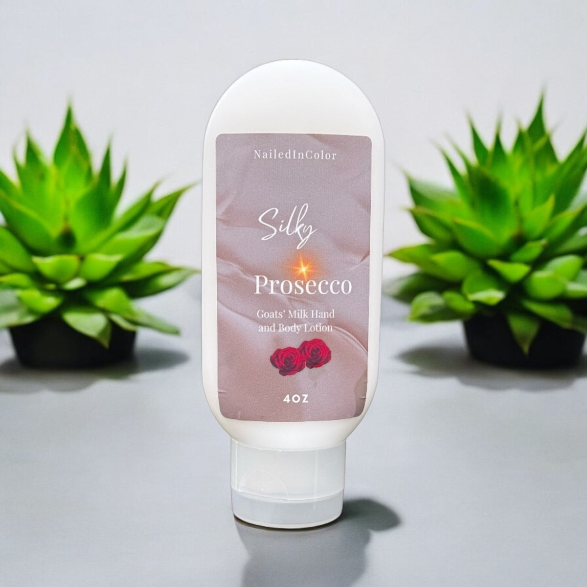 A white squeeze bottle of NailedInColor Silk Prosecco hand and body lotion stands centered on a gray surface, with two green potted succulents blurred in the background.